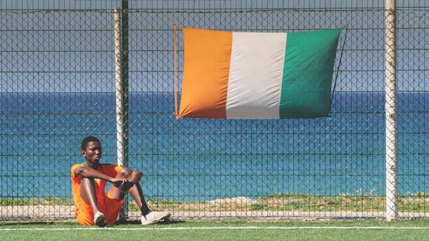 Young athlete in orange jersey sitting by an Ivorian flag on a football field with sea view.