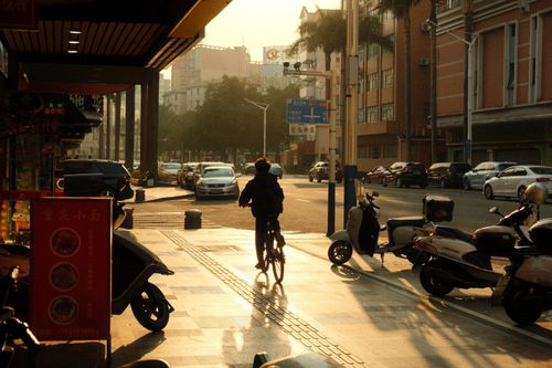 Free A cyclist rides through a sunlit city street lined with motorbikes and buildings at dusk. Stock Photo