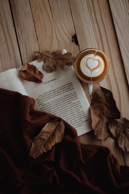 A latte with art next to an open book and autumn leaves on wooden surface.