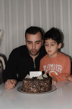 A father and son blowing out candles on a chocolate birthday cake indoors.