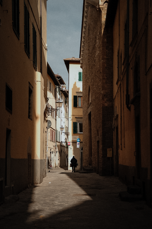 Free A narrow alleyway with old European architecture and a lone figure walking under the afternoon sun. Stock Photo