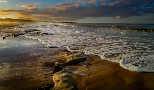 Free Tranquil beach scene capturing waves and rocks at sunrise, creating a peaceful ambiance. Stock Photo