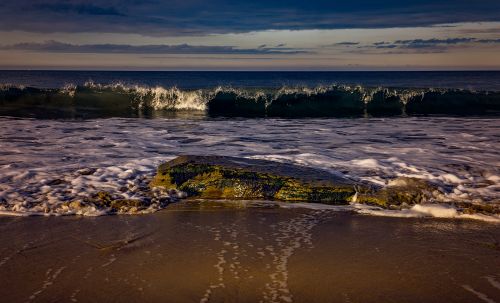 Free A tranquil beach scene with waves gently crashing on a rock at sunset. Stock Photo