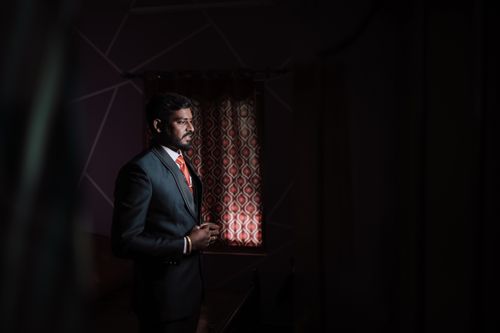 Free A portrait of a groom in formal attire standing by a patterned curtain in a dimly lit room. Stock Photo