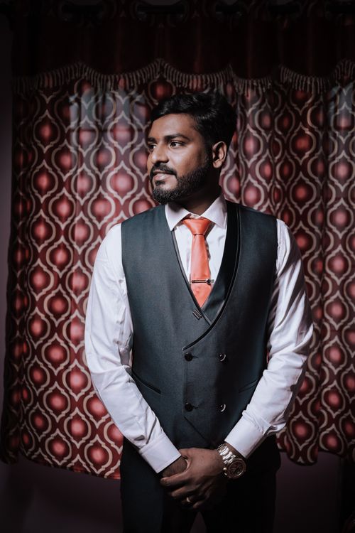 Free Stylish Indian groom in a dark suit and orange tie posing indoors with patterned curtains. Stock Photo