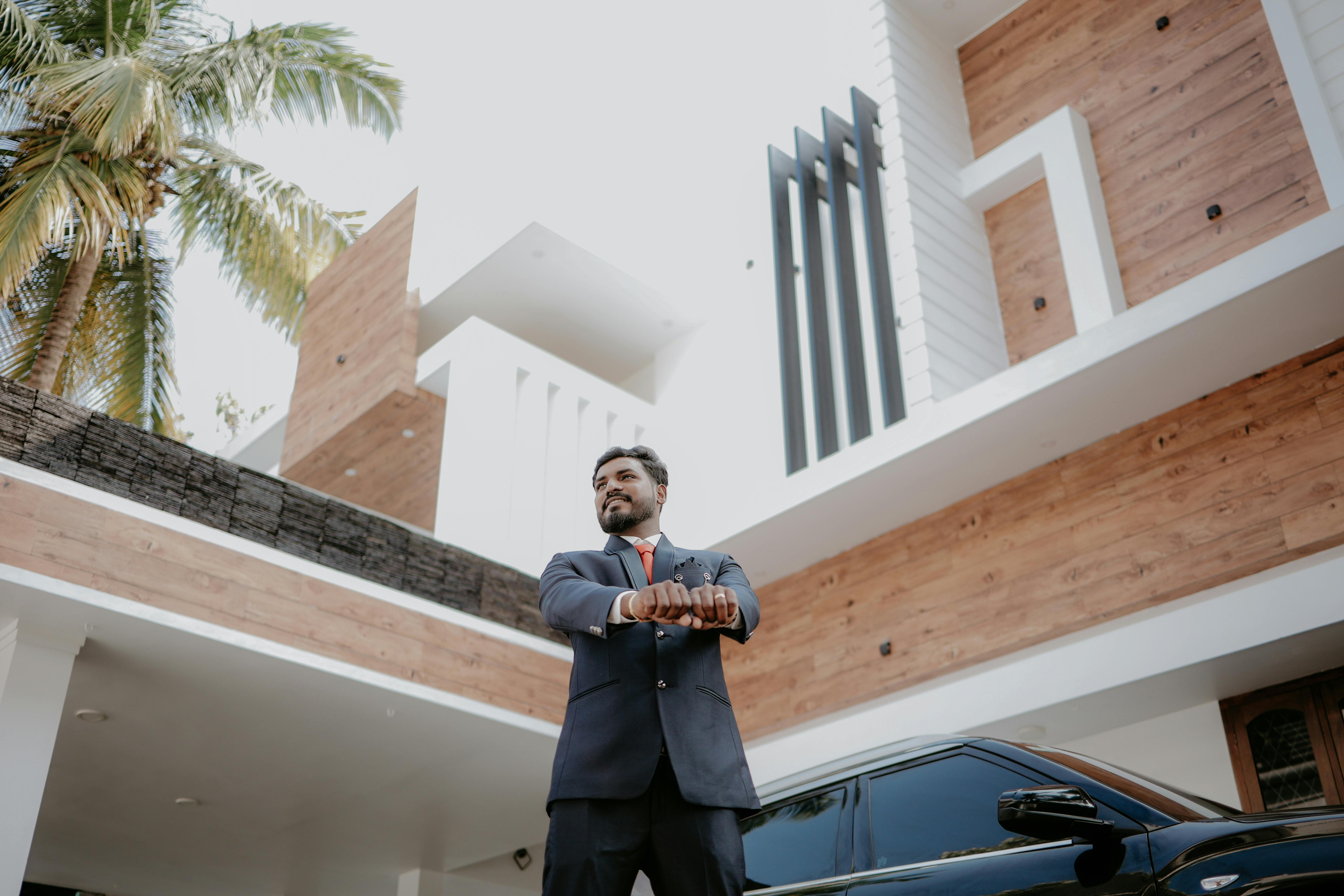A man in a suit confidently stands beside a black car and modern home exterior.