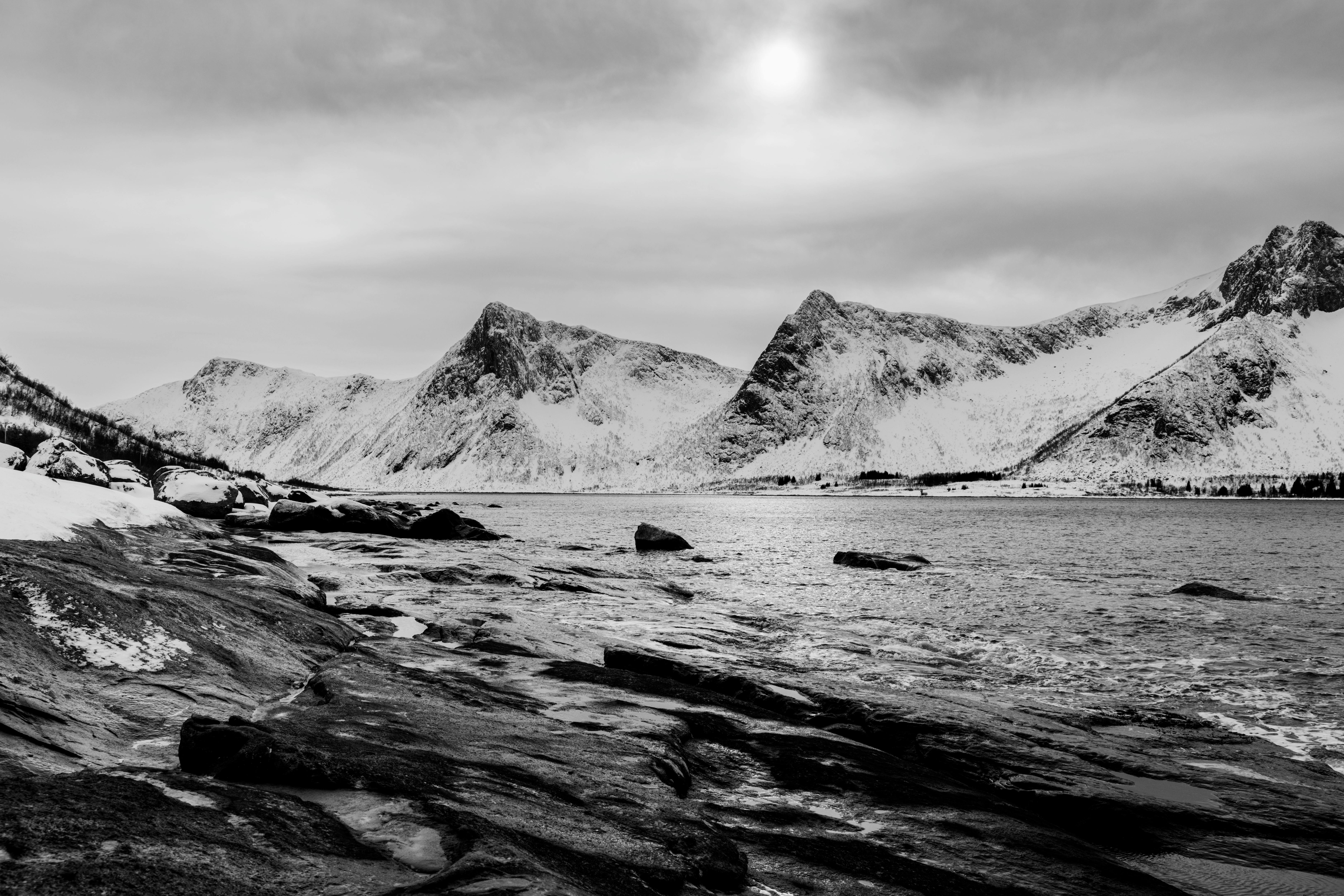 Stunning black and white image of snowy mountains and a rocky shoreline during winter.