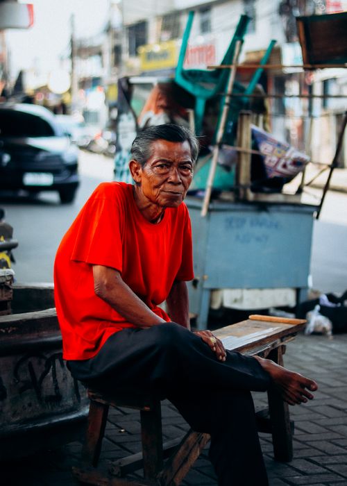 Free Elderly man in a vibrant red shirt sitting on a street in Jakarta market, Indonesia. Stock Photo