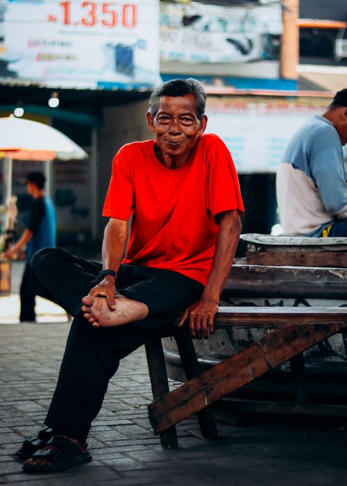 Free Elderly man in a red shirt sitting at a Jakarta market, smiling warmly. Stock Photo