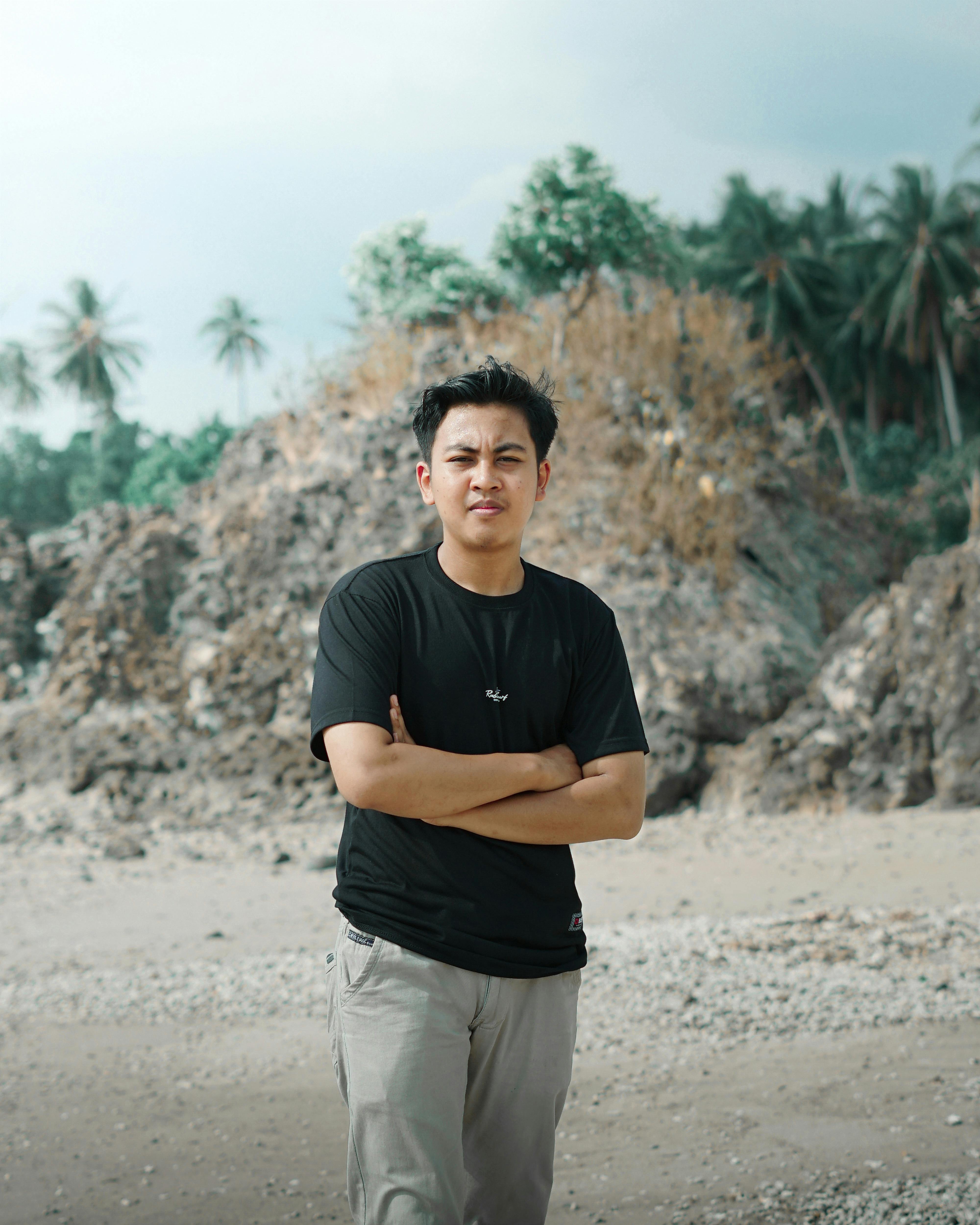 Portrait of a young man in casual attire standing on a rocky beach with tropical foliage.