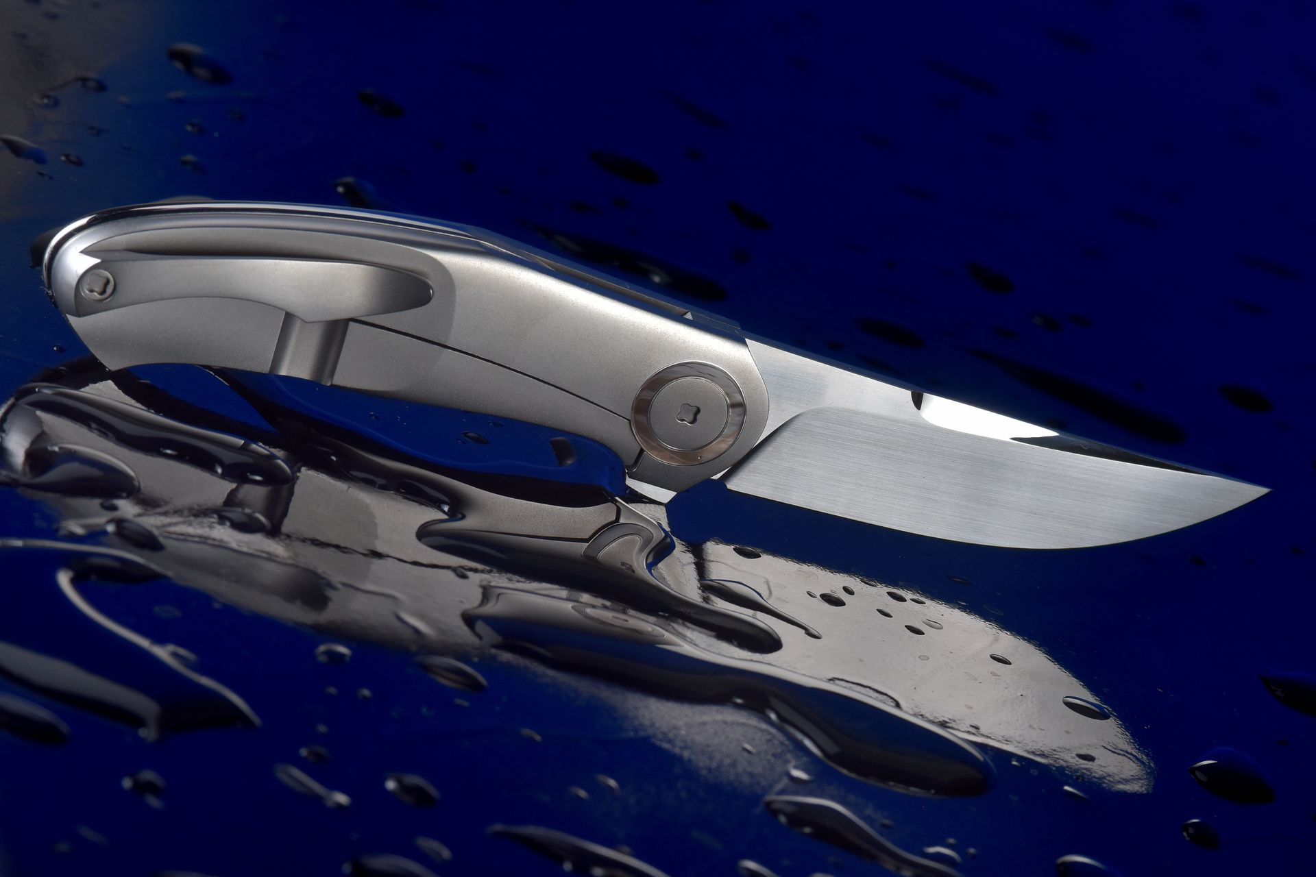 Close-up of a metallic folding knife against a sleek reflective surface with water droplets.