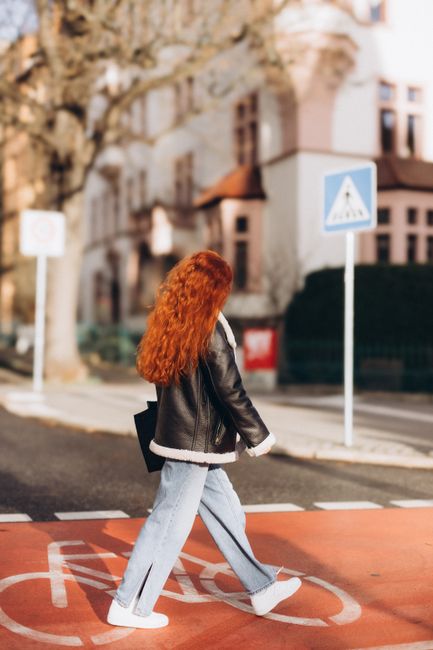 Red-haired woman in jacket crossing a city street on a bicycle lane.