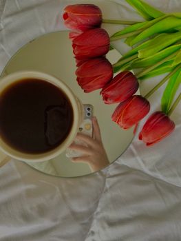 Flat lay of red tulips and coffee cup on a mirror, reflecting a hand with a phone, creating a cozy and artistic vibe.