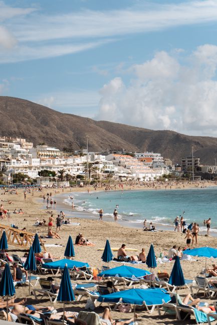 Vibrant beach scene on Canary Islands with sunbathers, waves, and mountain backdrop.
