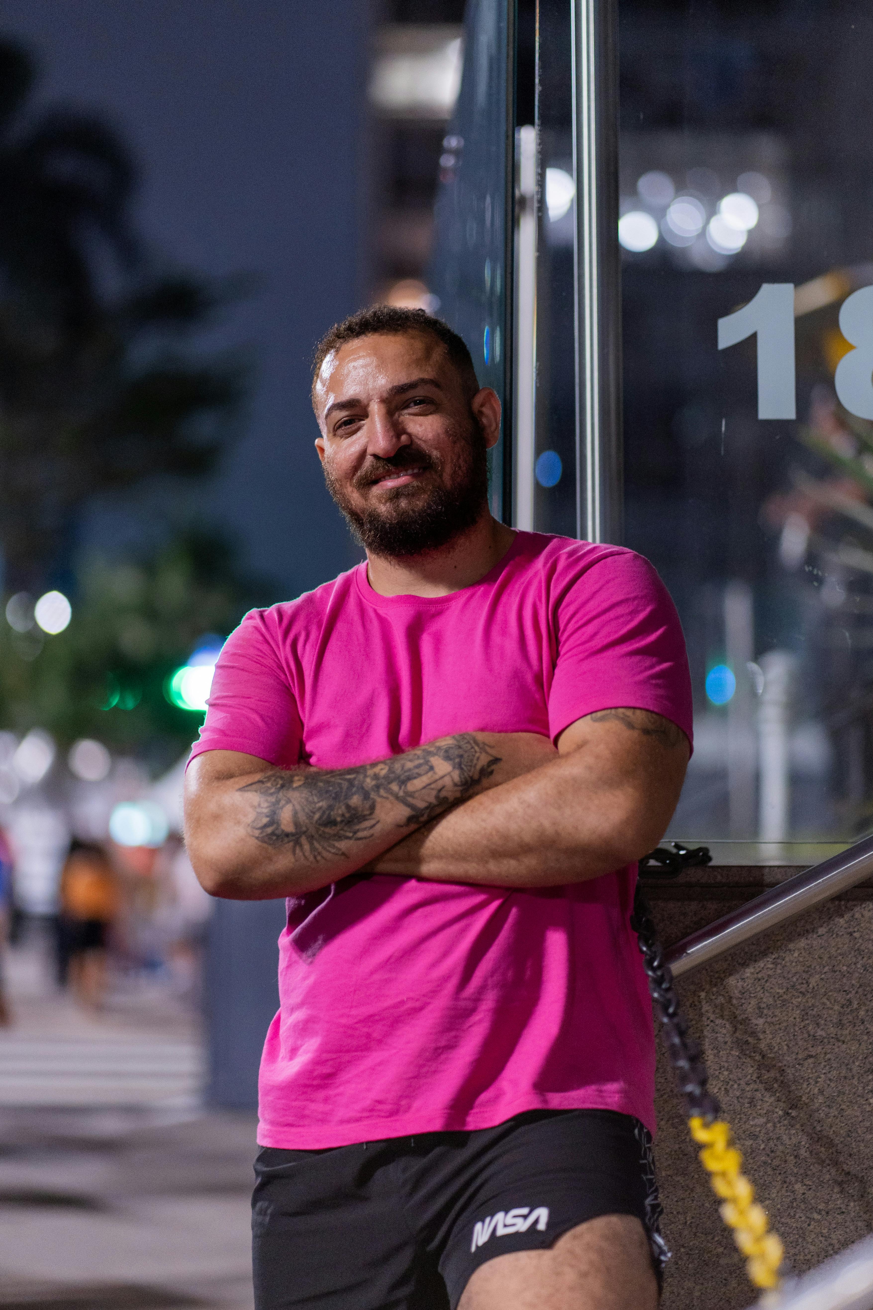 Bearded man in pink shirt leans against building, city night