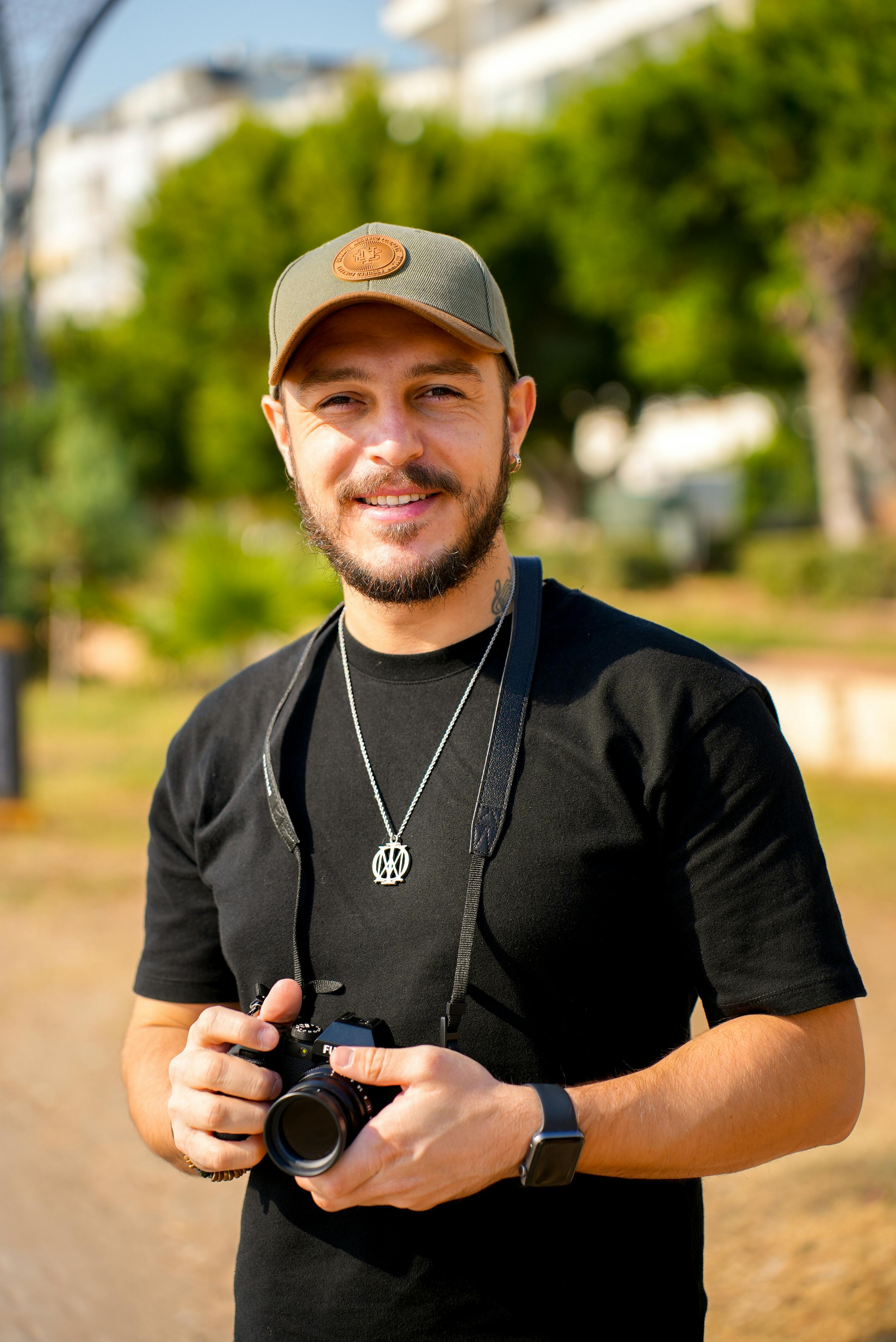 Smiling man holding a camera outdoors in a sunny park setting.