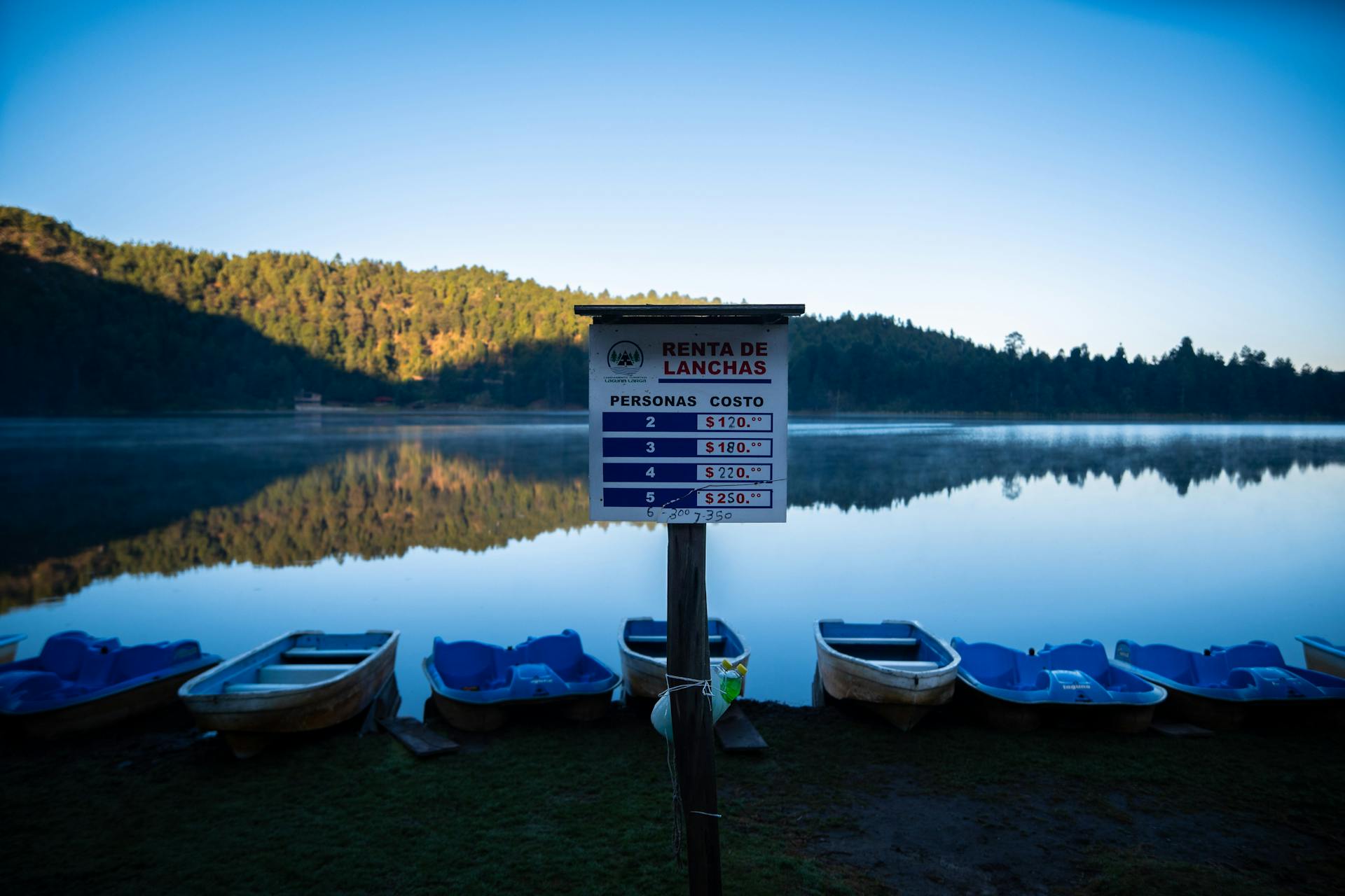 Tranquil morning at Los Azufres lake with boat rental sign and forest reflection.