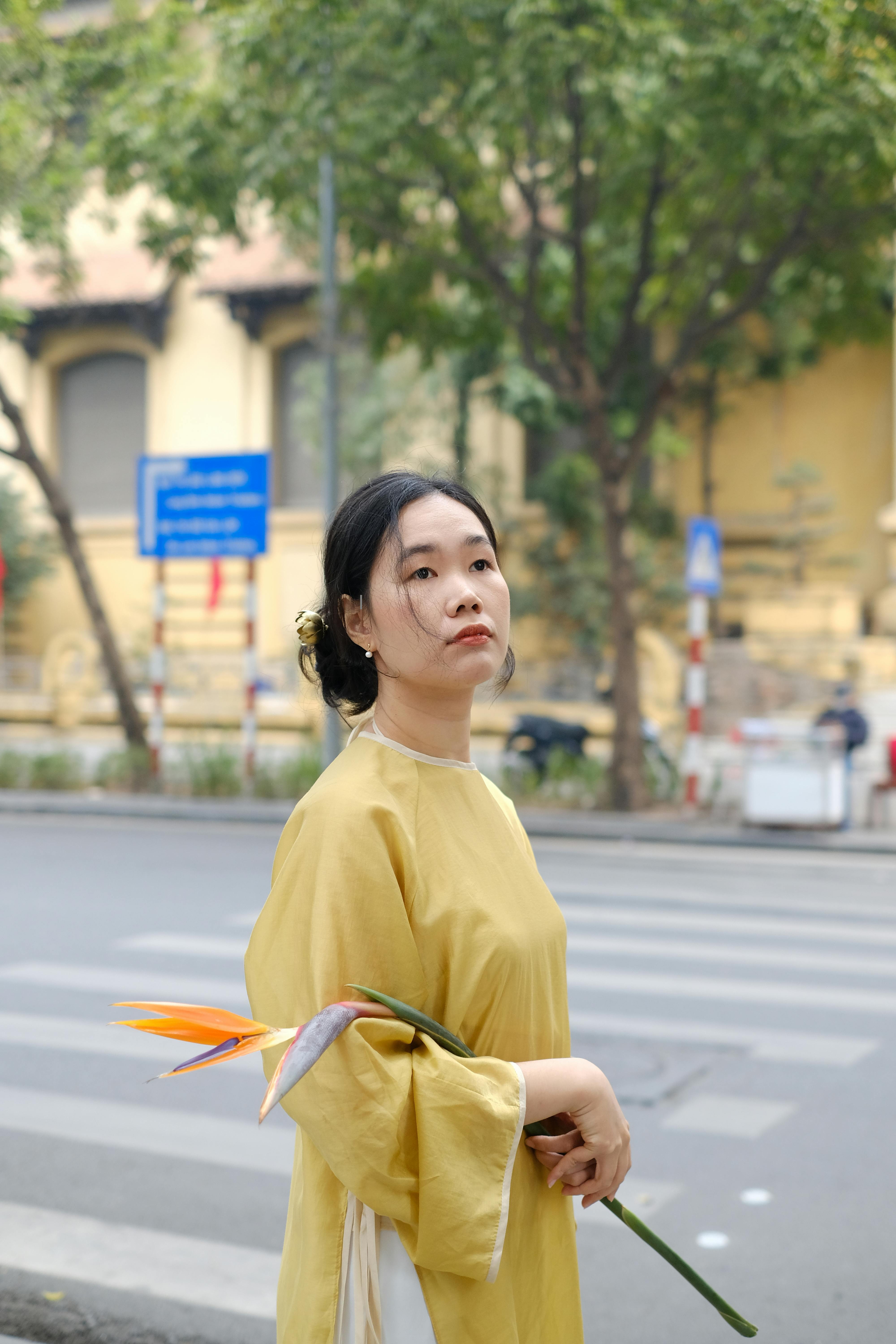 A woman in a yellow ao dai holds a flower while standing on a city street.