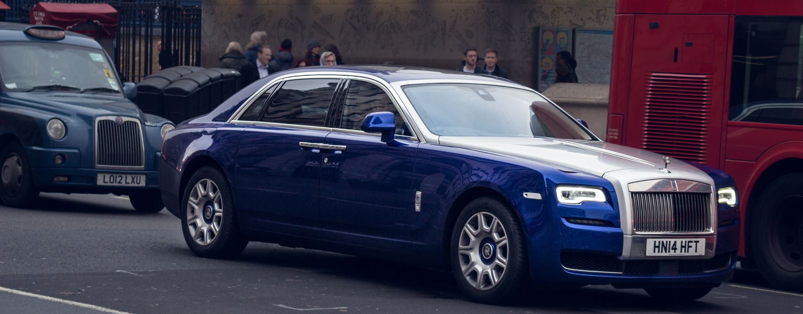 A blue luxury car driving on a busy street in London, surrounded by iconic vehicles.