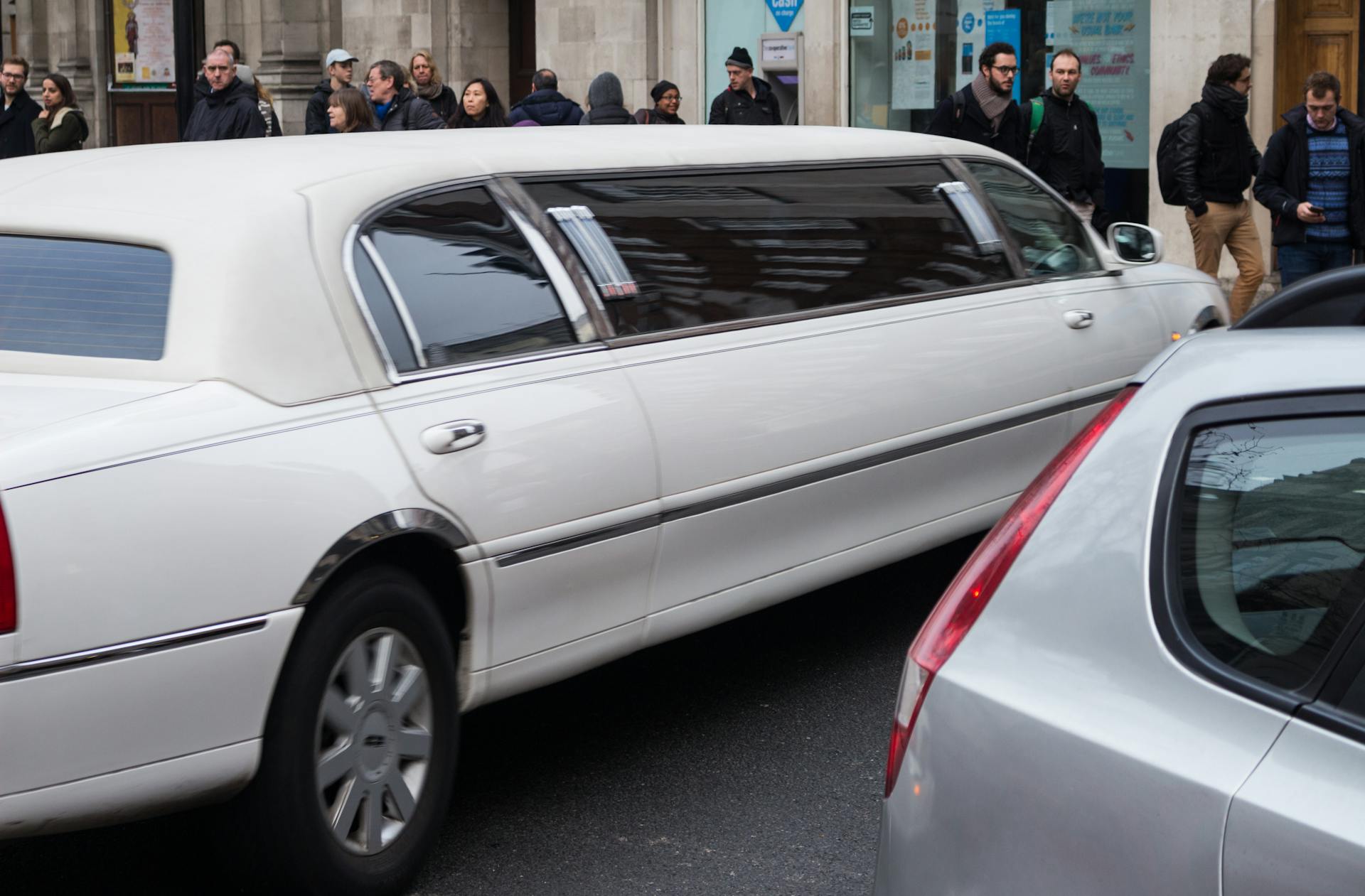 A white limousine driving down a bustling city street with pedestrians and other vehicles.