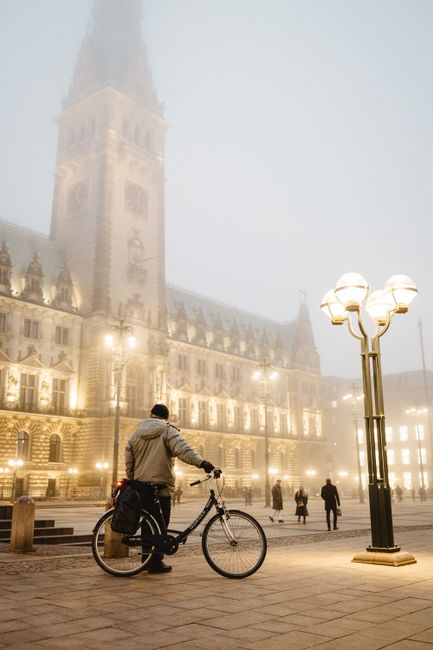 A lone cyclist stands with a bike in front of fog-shrouded Hamburg City Hall at night.