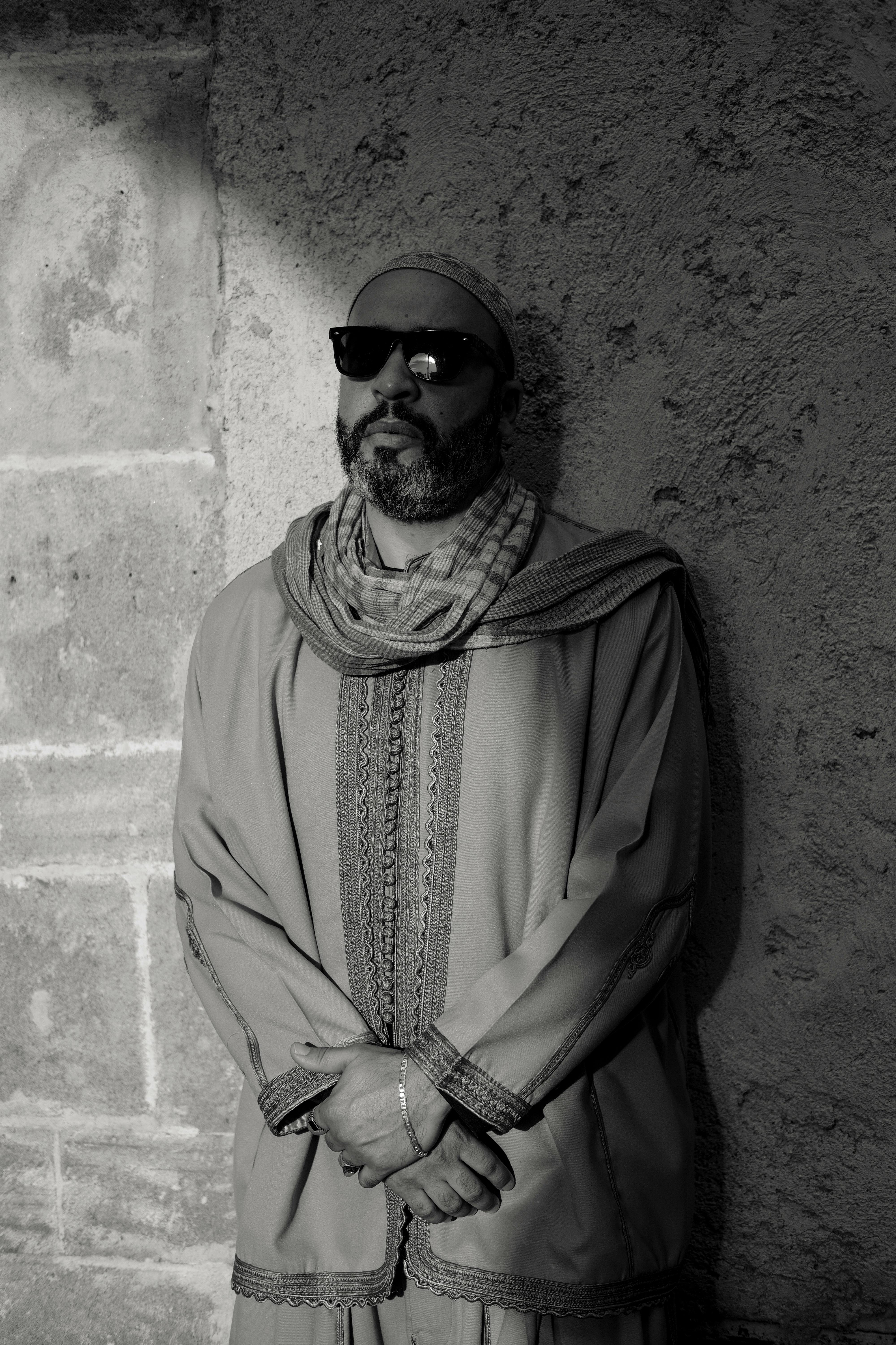 Black and white portrait of a man in sunglasses and traditional Moroccan attire against a stone wall.