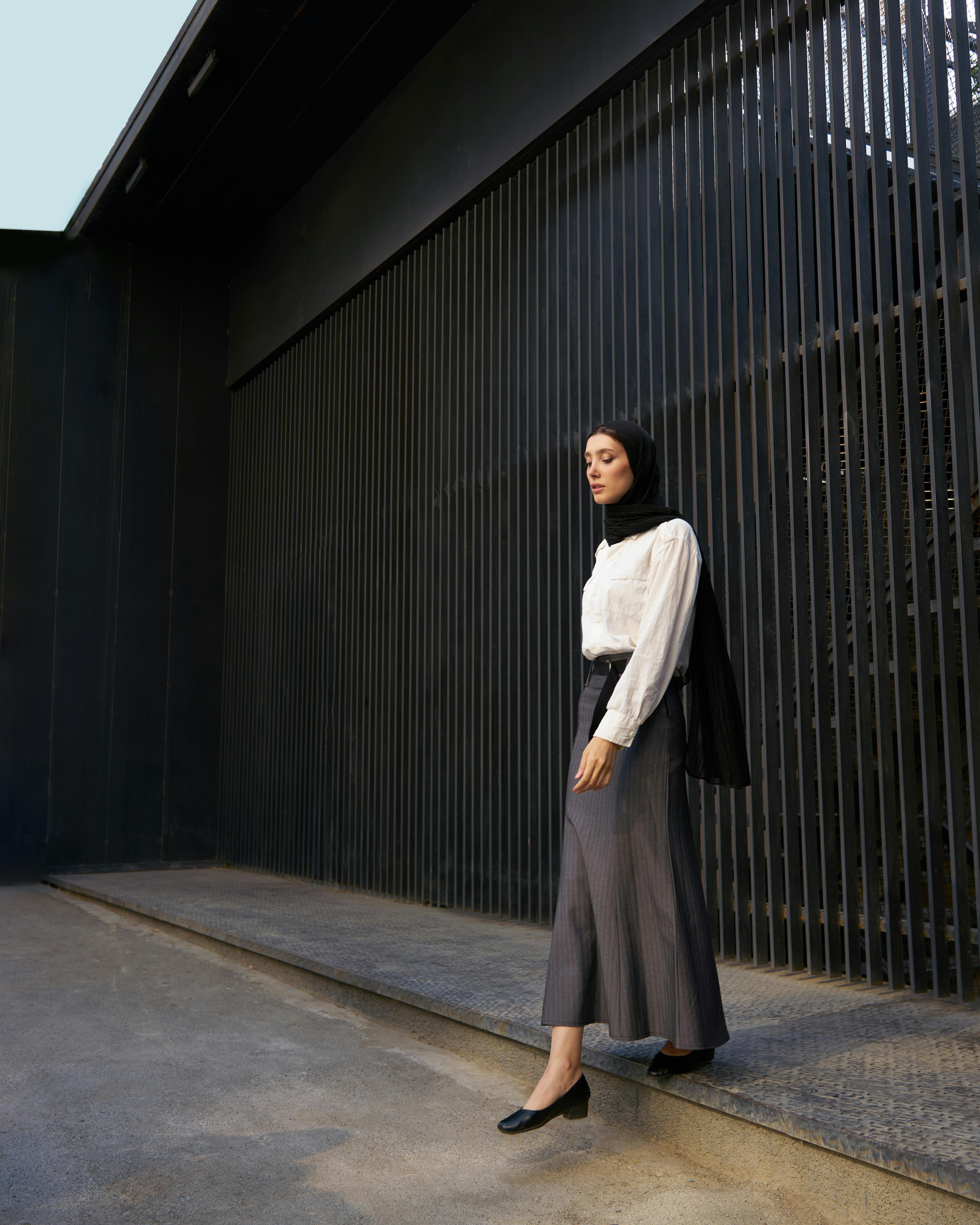 Stylish woman in hijab and modern attire walking outdoors by metal fence.