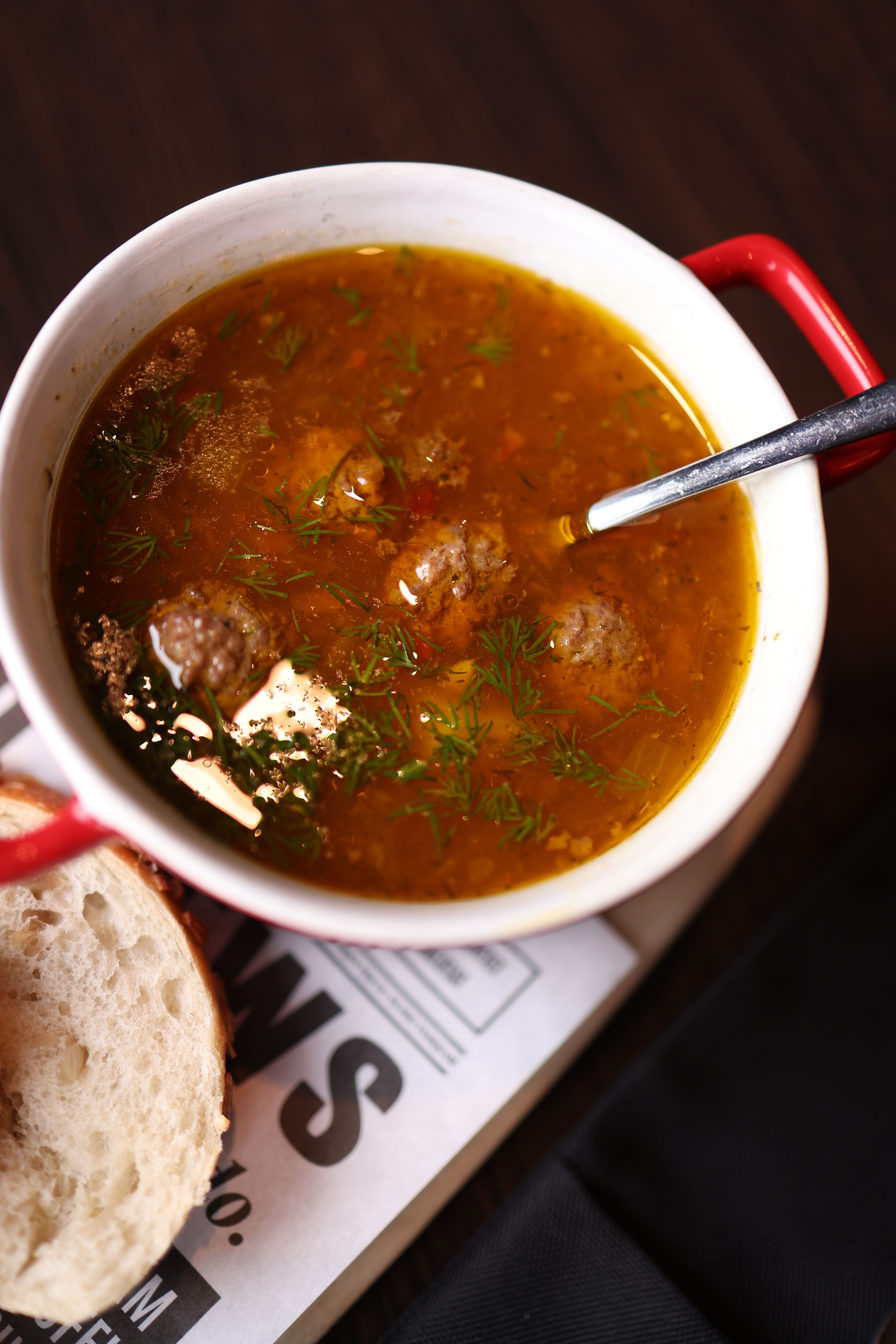 A bowl of savory meatball soup with herbs, served with fresh bread on a wooden table.