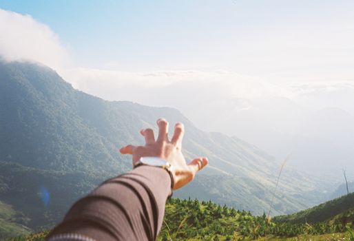 A hand stretches toward a stunning mountain range under a clear sky, capturing freedom.