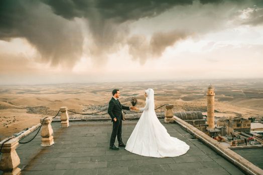 Elegant couple in wedding attire on an ancient rooftop with dramatic sky and landscape view.