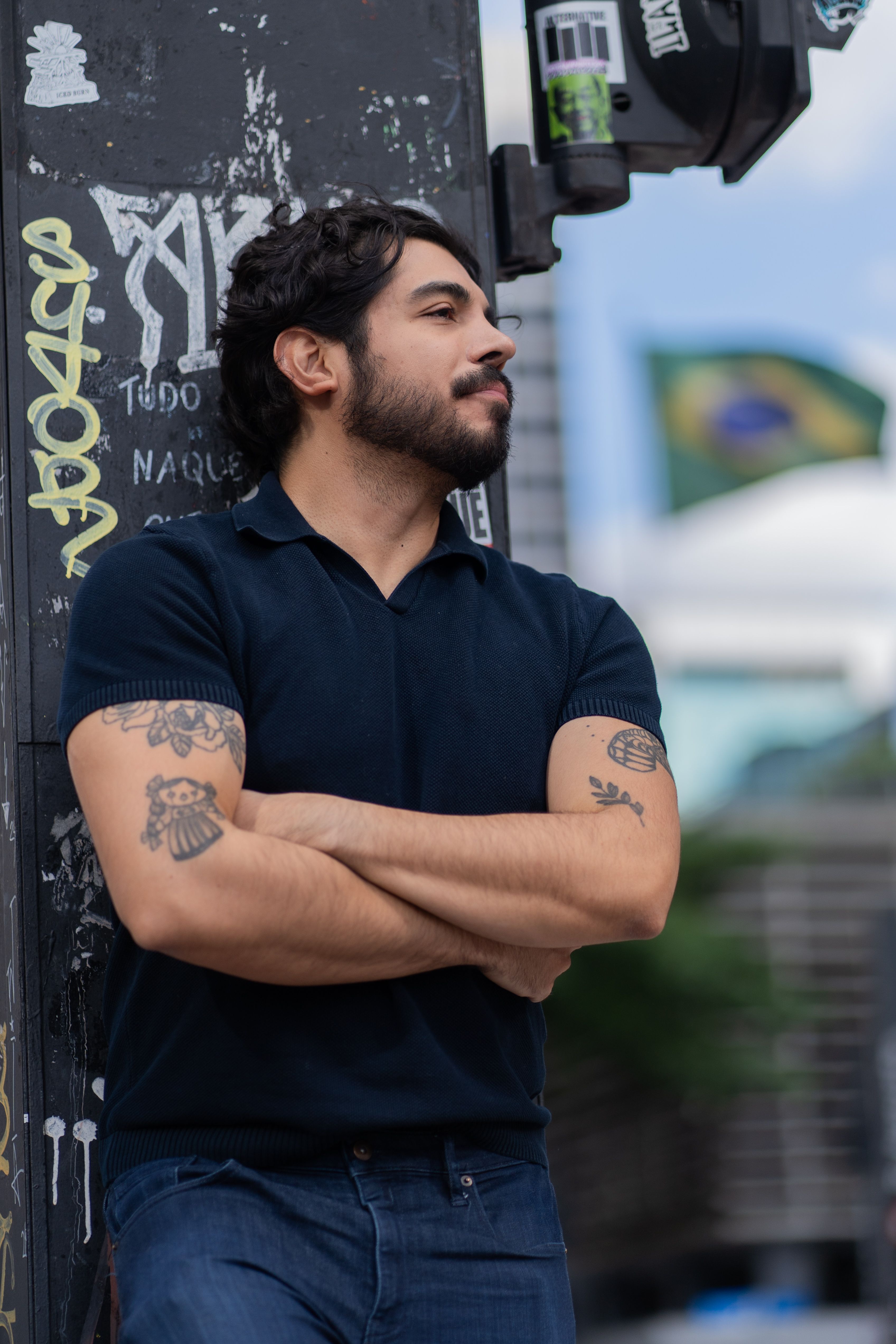 Man with tattoos confidently leans against a wall on an urban street, Brazilian flag in background.