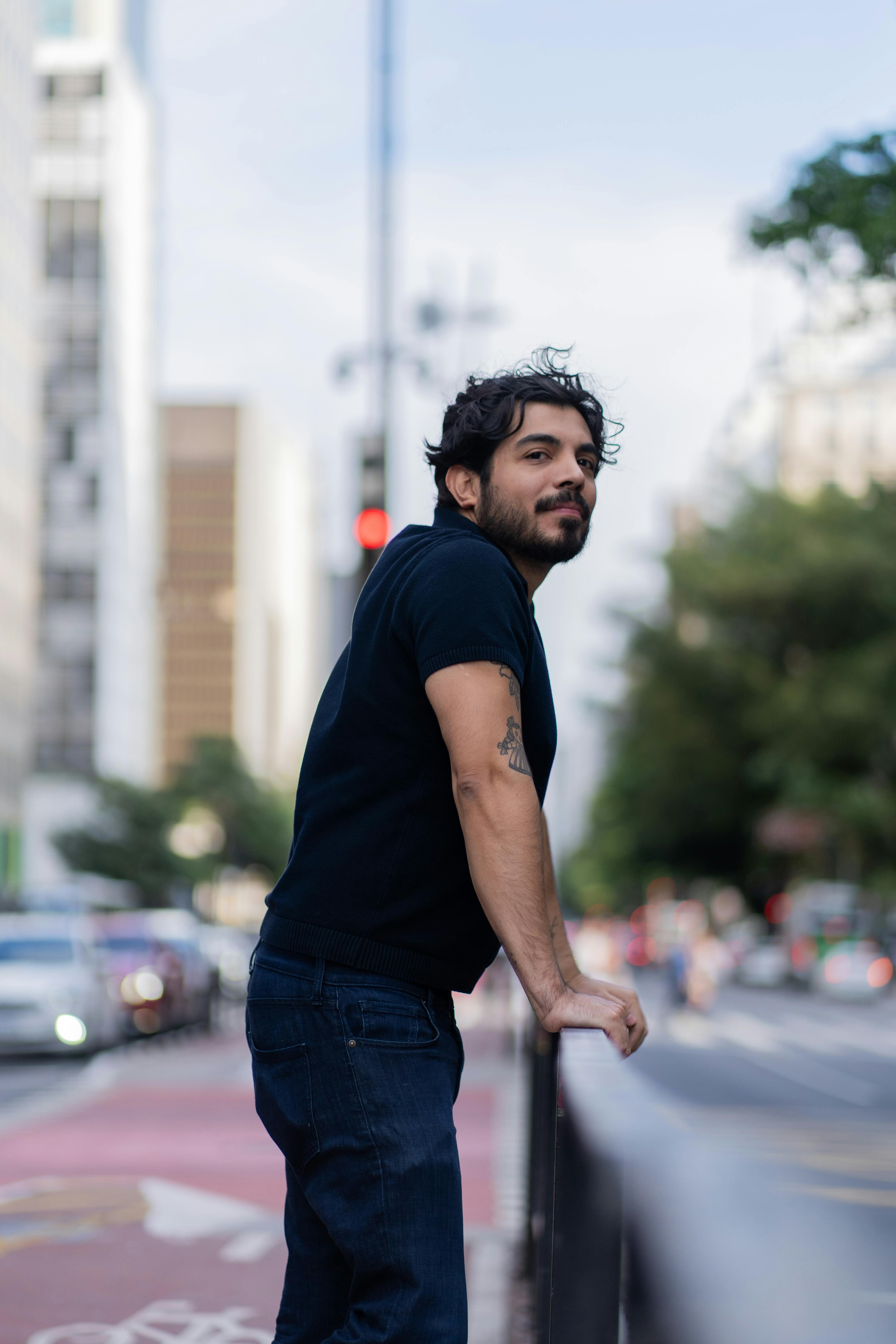 Young man leaning on a railing on a busy city street with trees and buildings in the background.