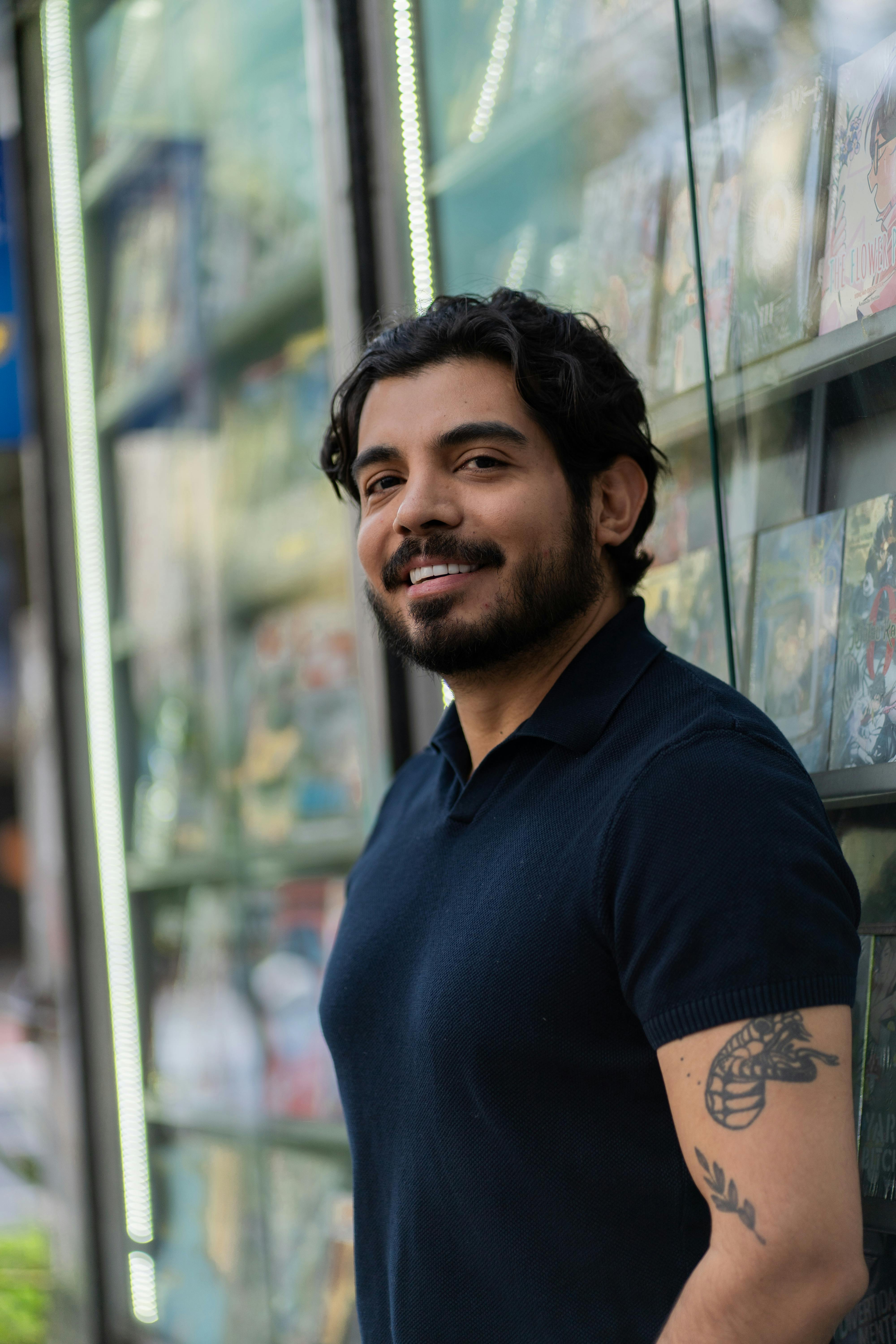 A bearded man with tattoos smiling near a storefront, casual attire.