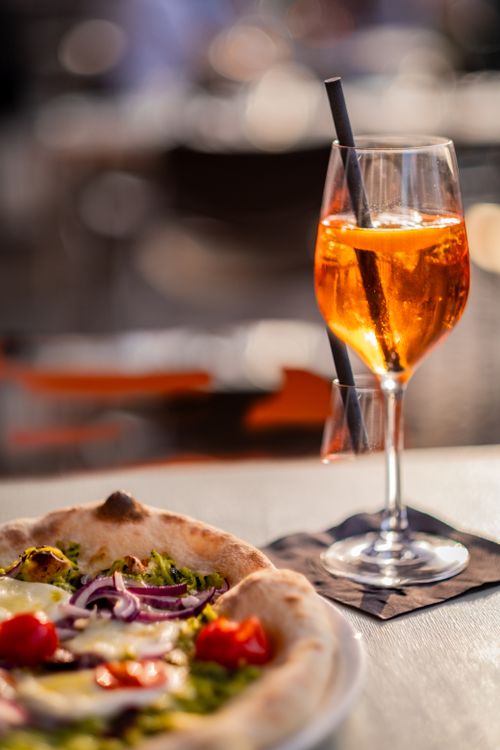Free A close-up of an Aperol Spritz cocktail next to a delicious gourmet pizza with blurred background. Stock Photo