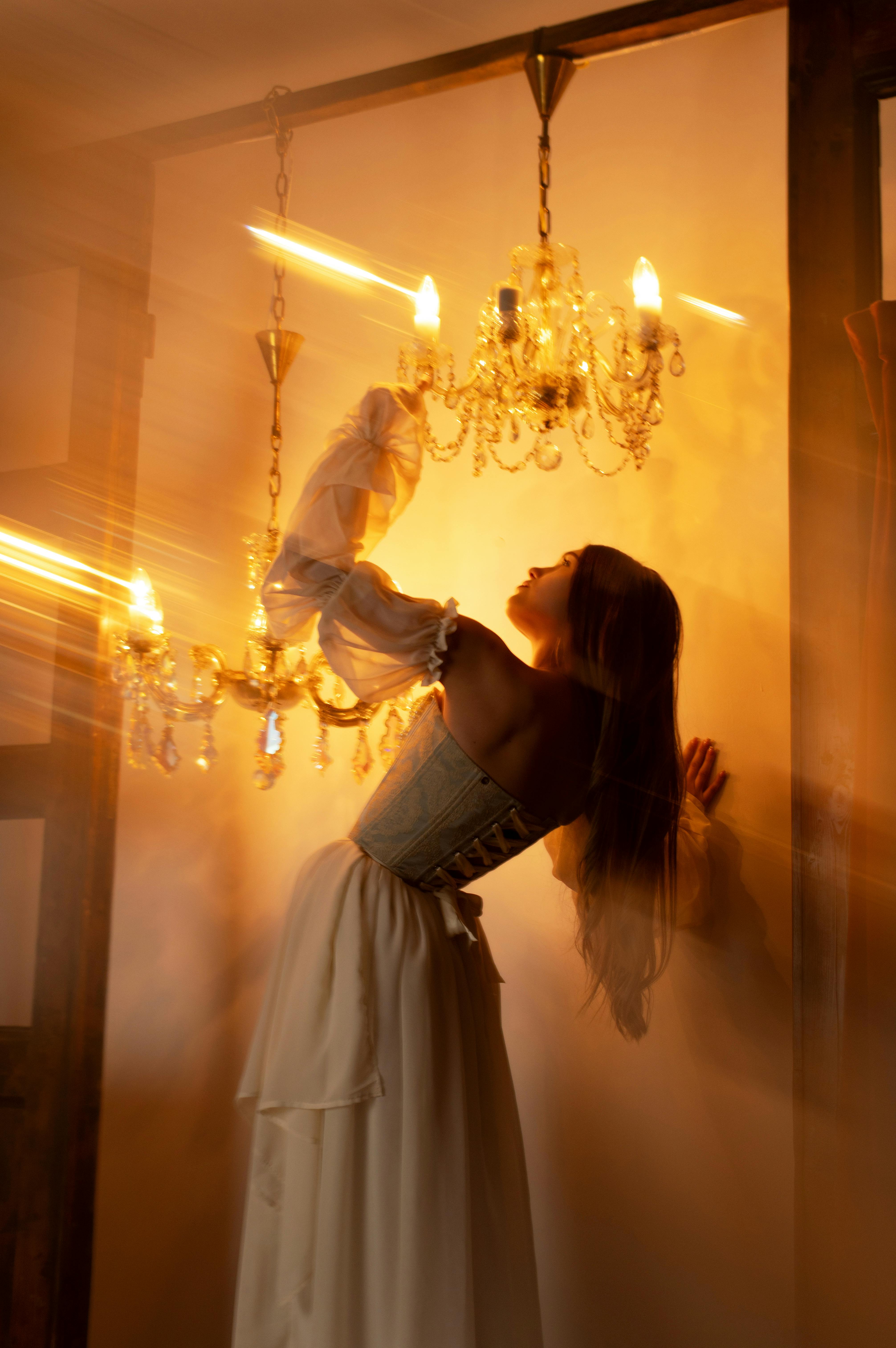 Elegant young woman in historical attire poses by a glowing chandelier in a warm, ambient setting.