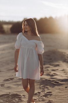 Woman in white dress walks barefoot on a beach at sunset, capturing serene summer vibes.