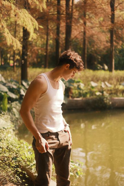 A young man in a white tank top stands by a serene forest pond, basking in sunlight.