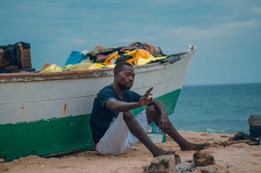 A man sits beside a colorful fishing boat on a sandy beach, enjoying a peaceful day.