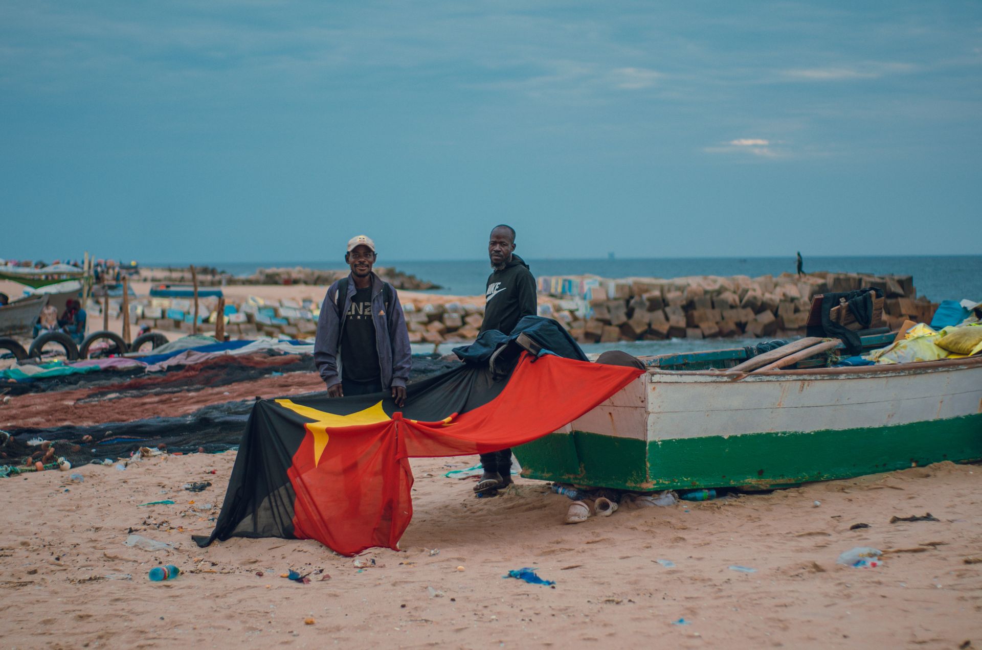 Two fishermen stand by a boat on a beach, holding the Angolan flag, under a cloudy sky.