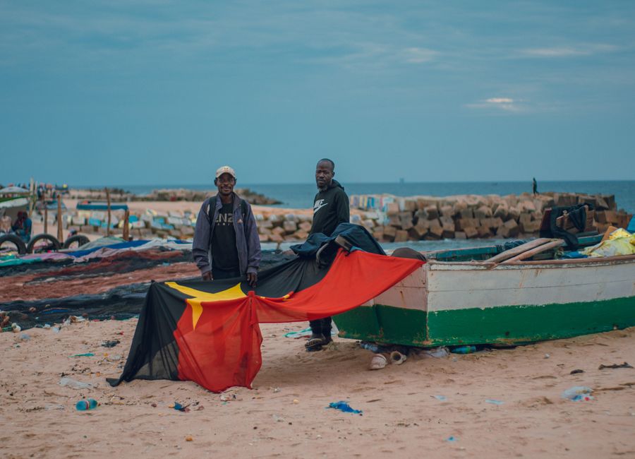 Two fishermen stand by a boat on a beach, holding the Angolan flag, under a cloudy sky.