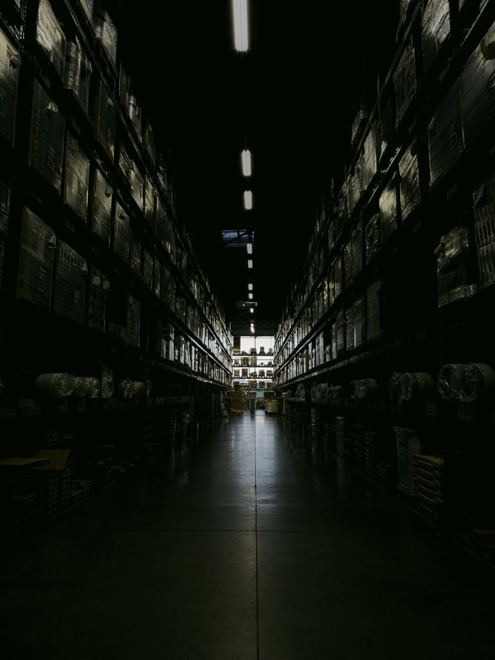 A dark, narrow warehouse aisle with shelves filled with boxes and goods, illuminated by overhead lights.