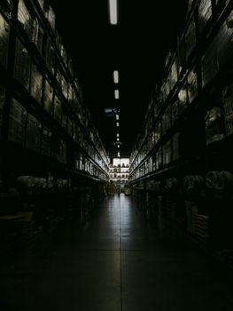 A dark, narrow warehouse aisle with shelves filled with boxes and goods, illuminated by overhead lights.