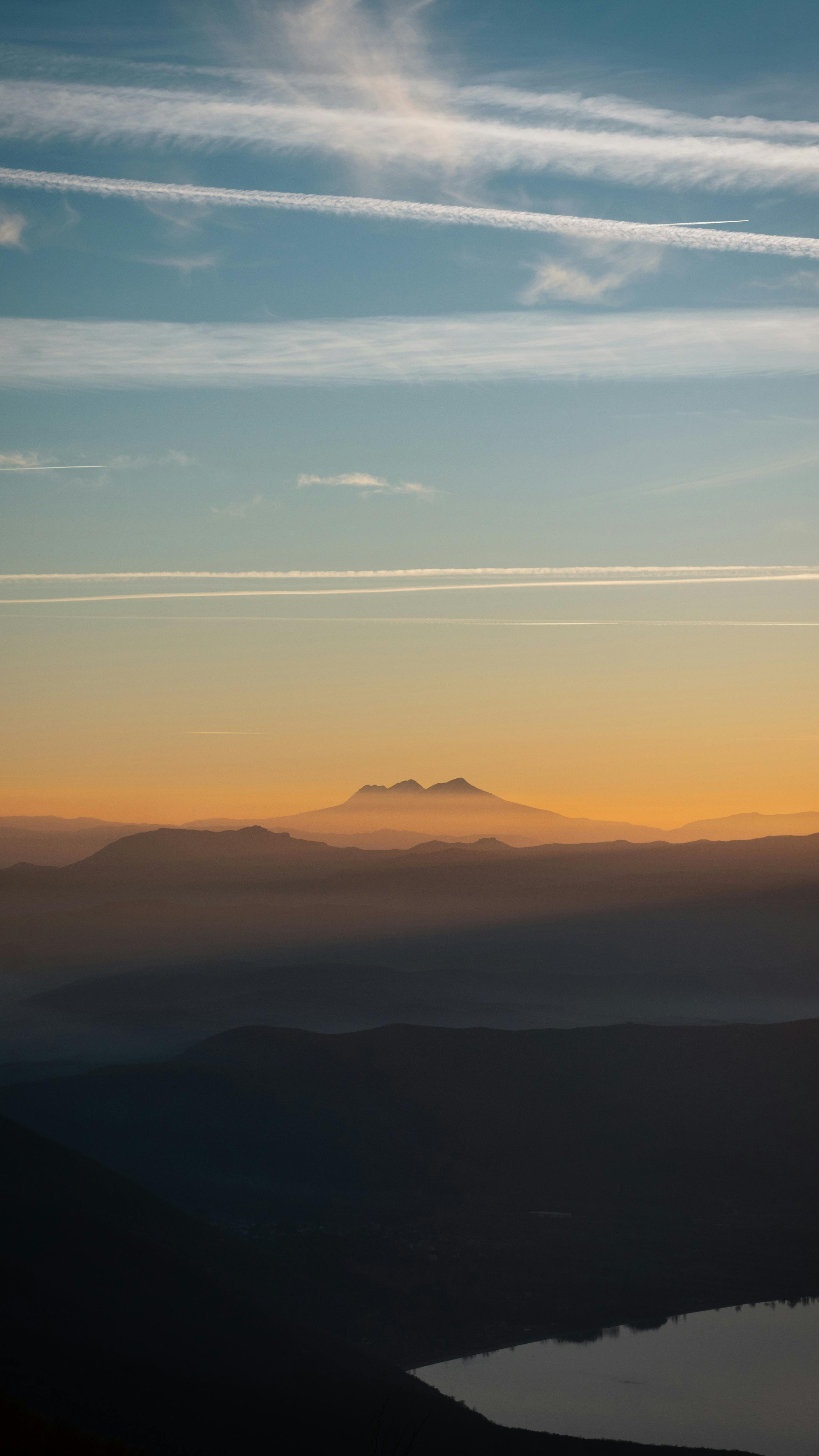 A tranquil sunset scene over mountain ranges with a clear sky and contrails.