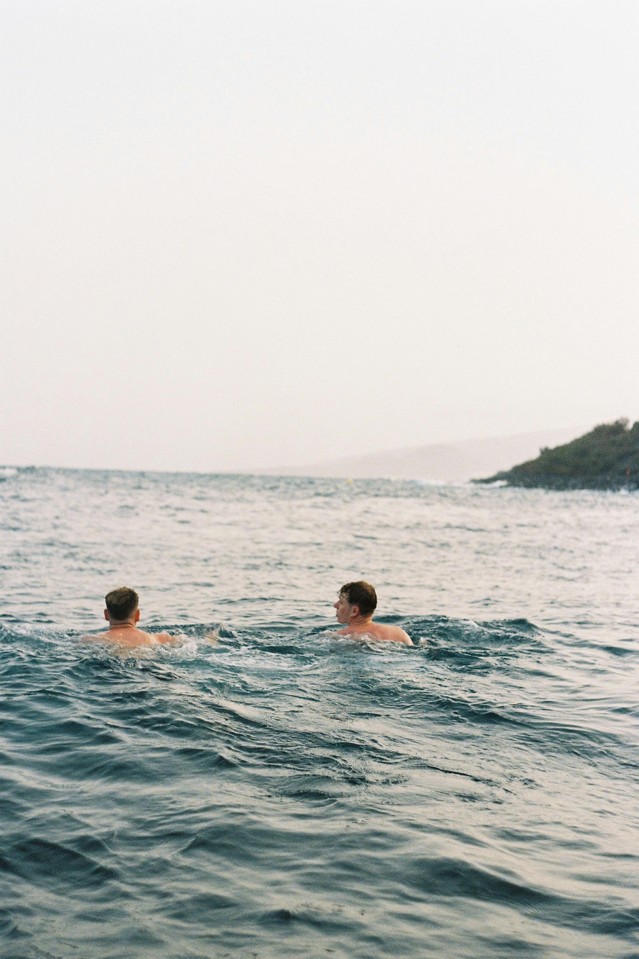 Two men swimming in a calm ocean near a rocky shore during dusk, creating a serene scene.