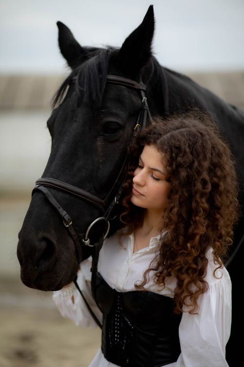 Free A young woman with curly hair leans affectionately against a black horse outdoors, conveying a serene connection. Stock Photo