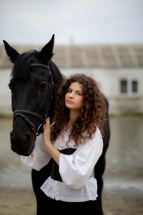 Free Curly-haired woman in vintage attire posing with a black horse outdoors. Stock Photo