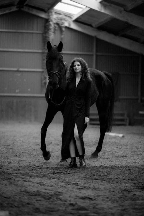 Free A woman in a black dress stands confidently with a black horse in an indoor equestrian arena. Stock Photo