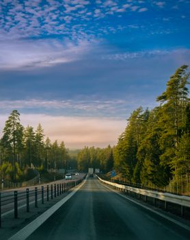 A serene highway through a lush pine forest under a vibrant sunrise sky.