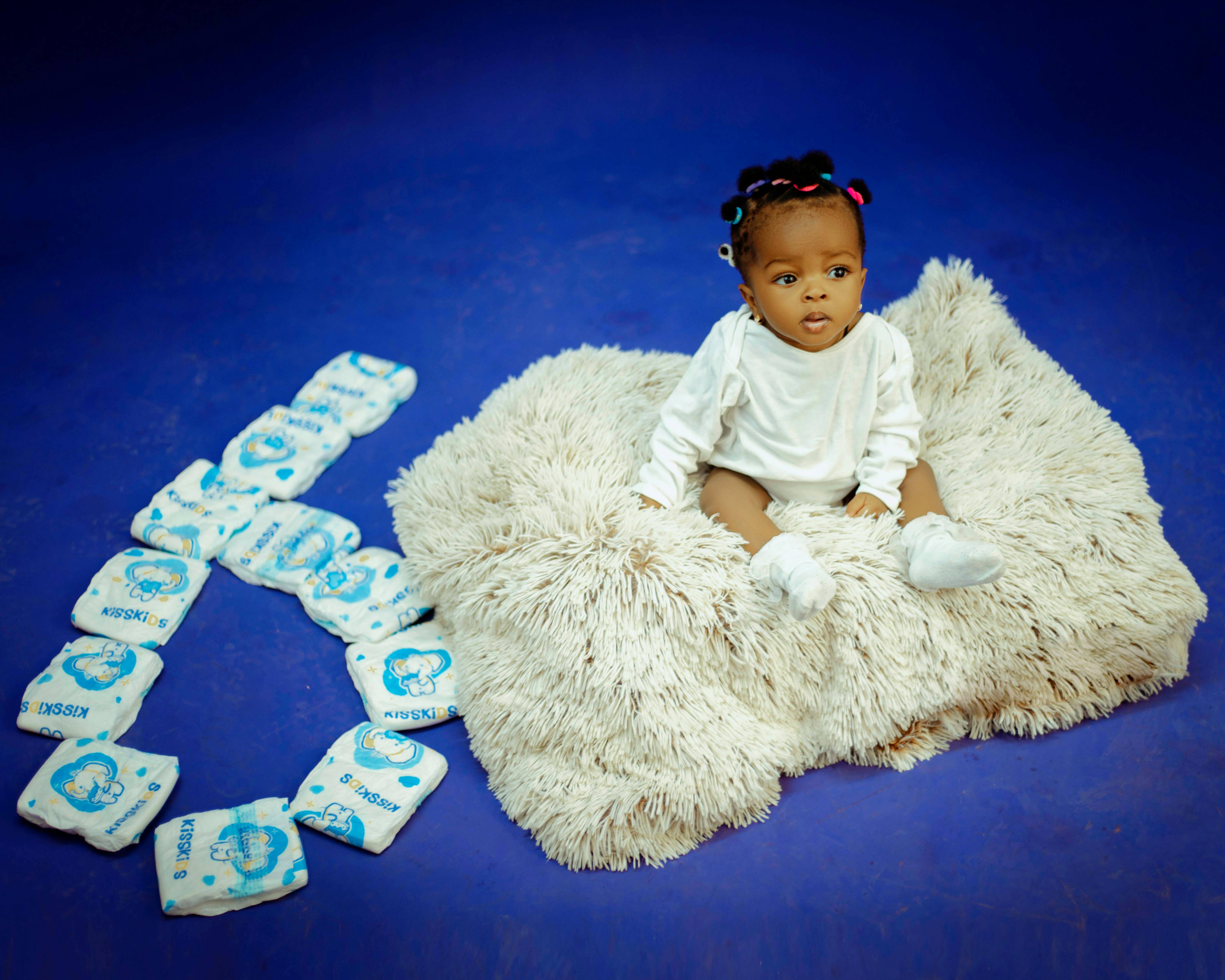 Adorable baby sitting on a fluffy rug surrounded by arranged diapers on a blue background.