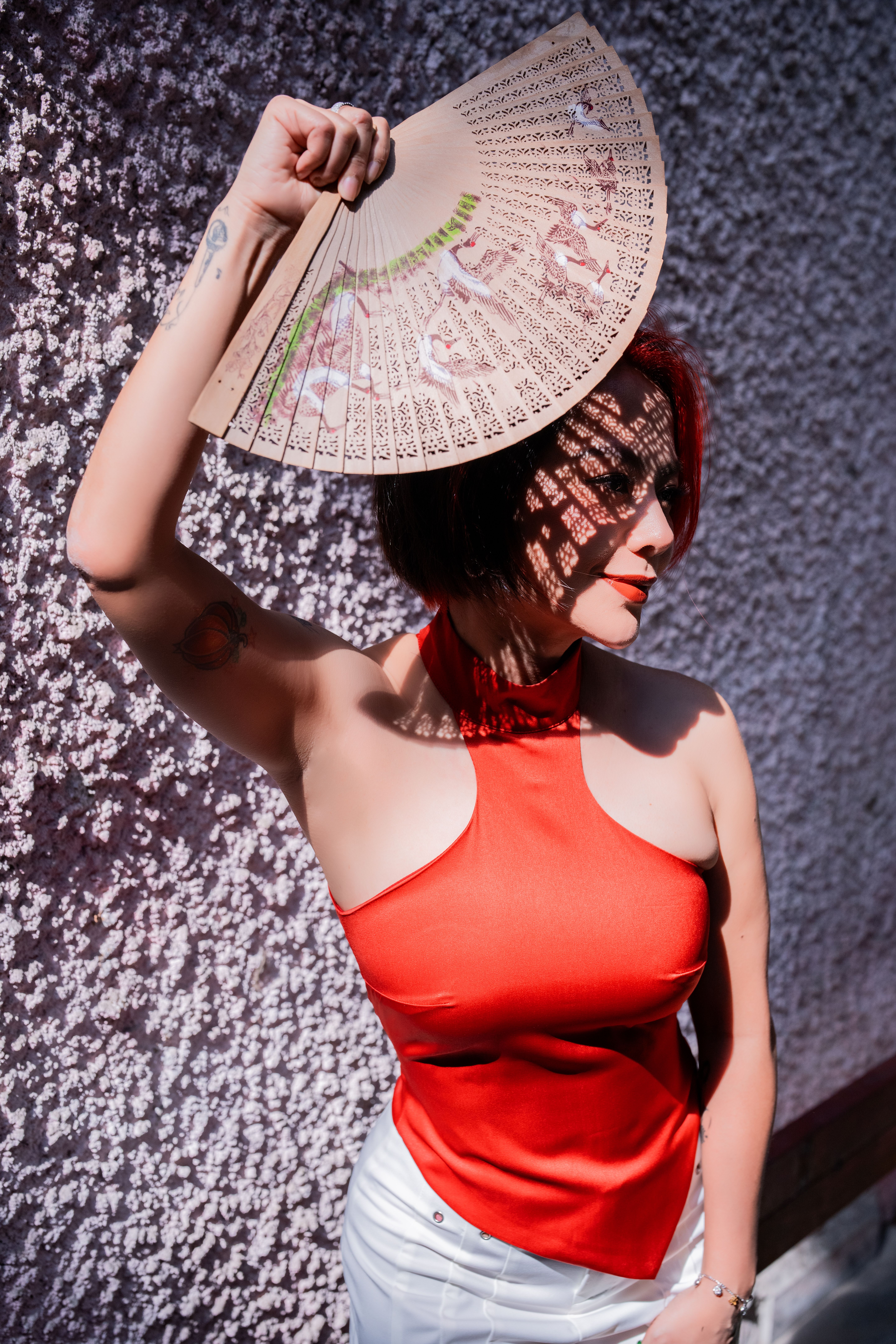 Stylish woman in red top with a fan enjoying sunlight against a textured wall.